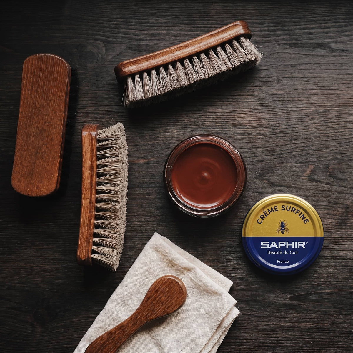 Crème Surfine Saphir Cognac en pot ouvert, avec des brosses en crin et une chamoisine, vue de dessus