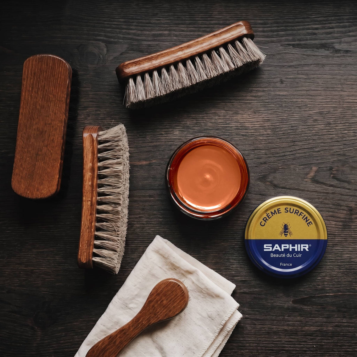 Crème Surfine Saphir Cognac clair en pot ouvert, avec des brosses en crin et une chamoisine, vue de dessus