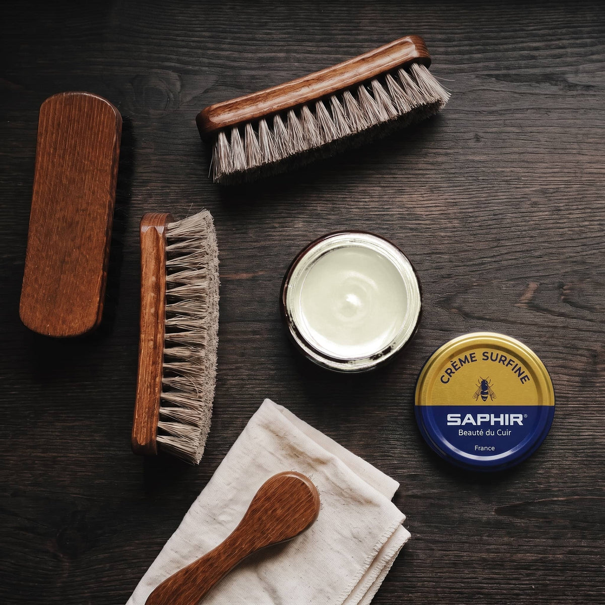 Crème Surfine Saphir Fumée en pot ouvert, avec des brosses en crin et une chamoisine, vue de dessus