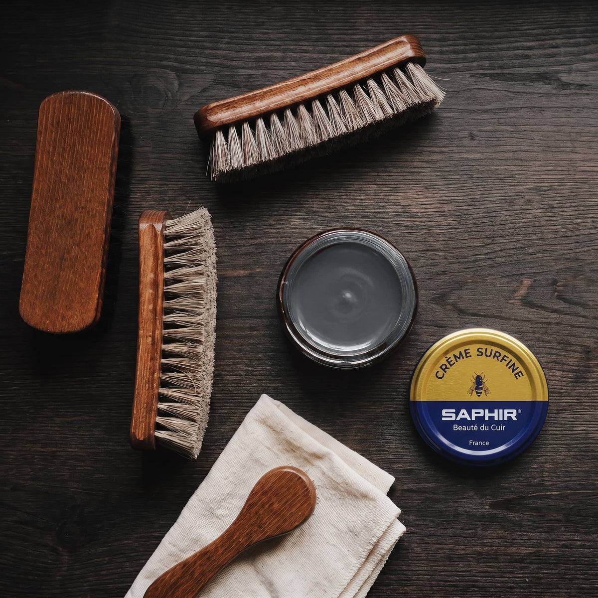 Crème Surfine Saphir Gris foncé en pot ouvert, avec des brosses en crin et une chamoisine, vue de dessus