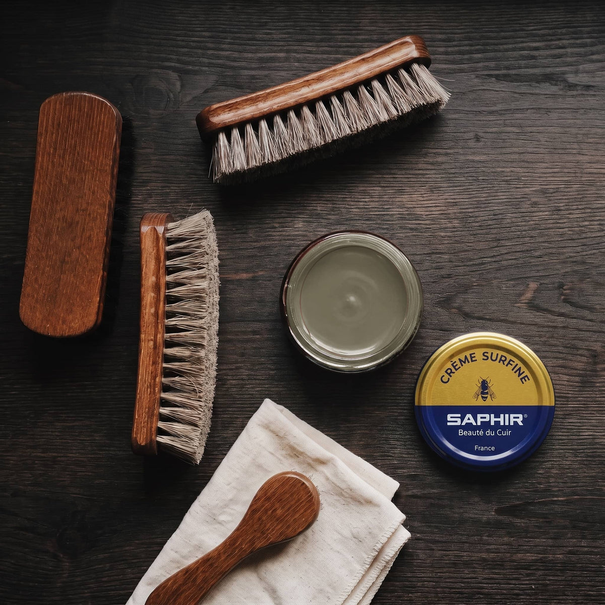 Crème Surfine Saphir Kaki en pot ouvert, avec des brosses en crin et une chamoisine, vue de dessus
