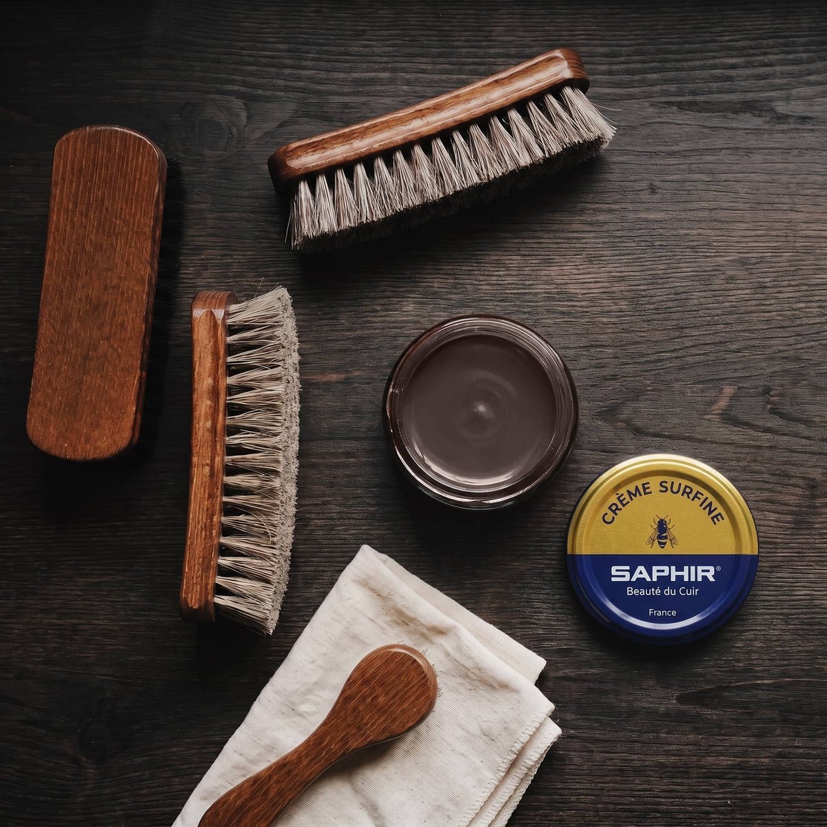 Crème Surfine Saphir Marron foncé en pot ouvert, avec des brosses en crin et une chamoisine, vue de dessus
