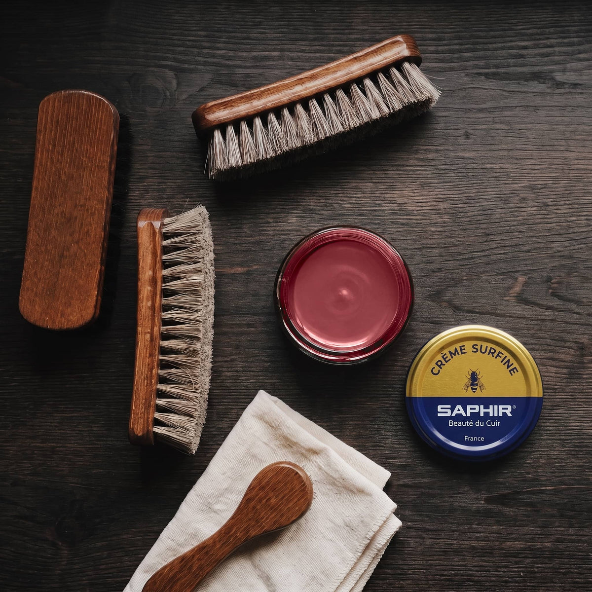 Crème Surfine Saphir Rouge Hermès en pot ouvert, avec des brosses en crin et une chamoisine, vue de dessus