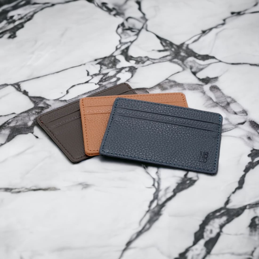 Les trois porte-cartes en cuir grainé, bleu marine, gold et marron foncé, sur un plateau de marbre